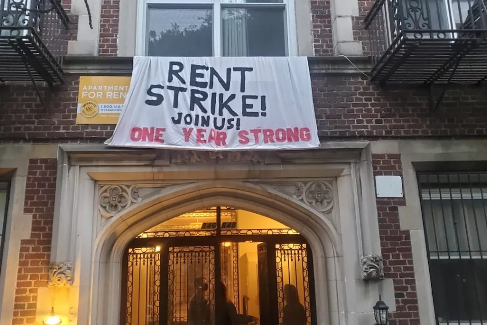 "Rent Strike - Join Us! One Year Strong" cloth banner hanging on red brick New York City apartment building.